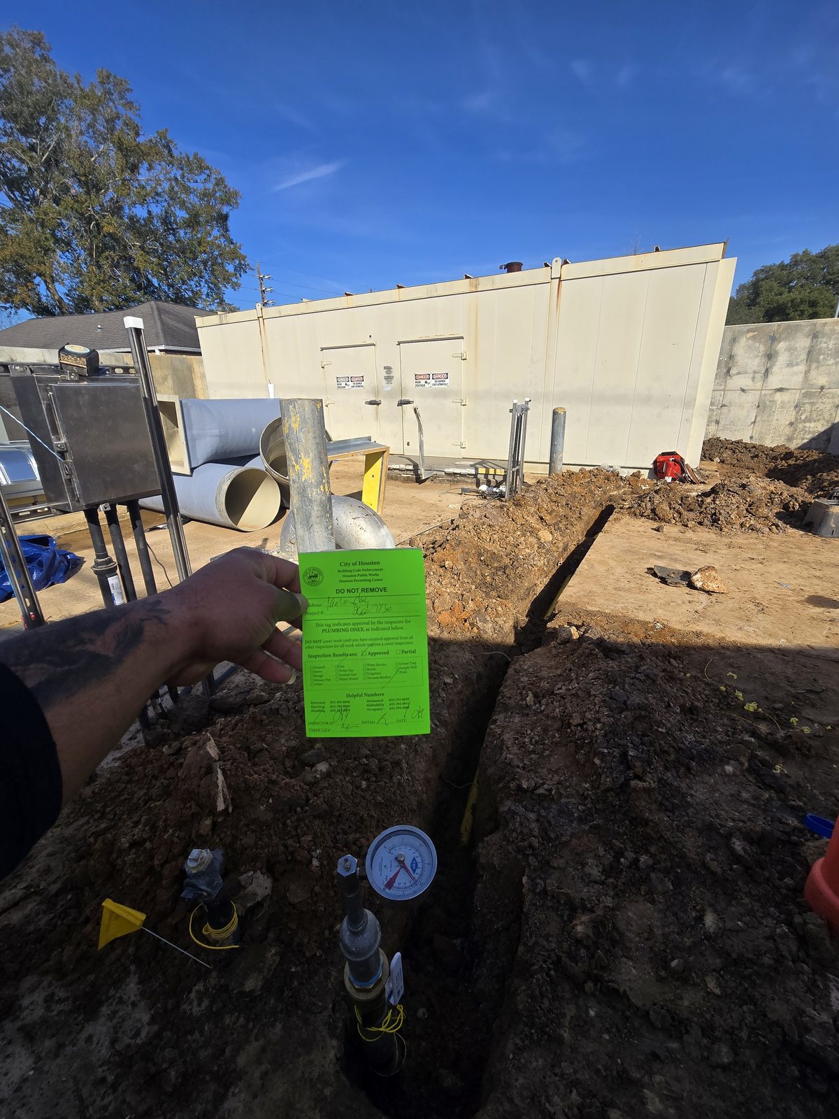 City of Houston plumbing inspection tag marked Approved, held over a completed trench on a commercial job site